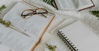 Open Bible with glasses, notepad, and pen on a white quilt, evoking study and reflection.