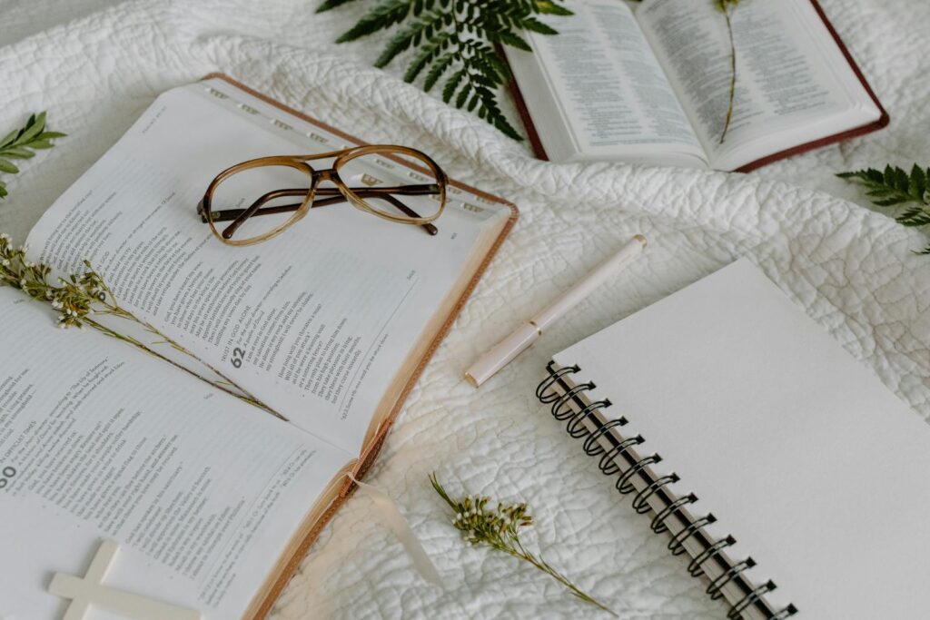 Open Bible with glasses, notepad, and pen on a white quilt, evoking study and reflection.